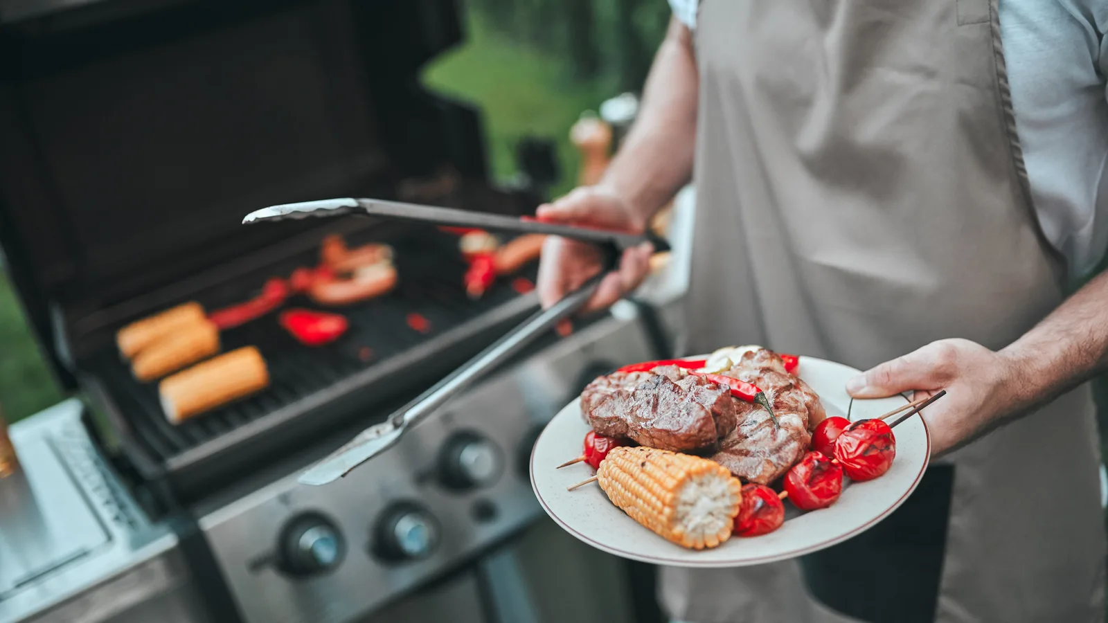 Man tending to the grill removing grilled corn, peppers and steak. He is wearing an apron and is hanging a plate towards the camera. New pet-friendly apartment homes for rent with private fenced backyards in Arizona, Texas, Florida, and North Carolina with pool, pickleball, dog park, gym, clubhouse and gated community. *No breed or weight restrictions. Yardly