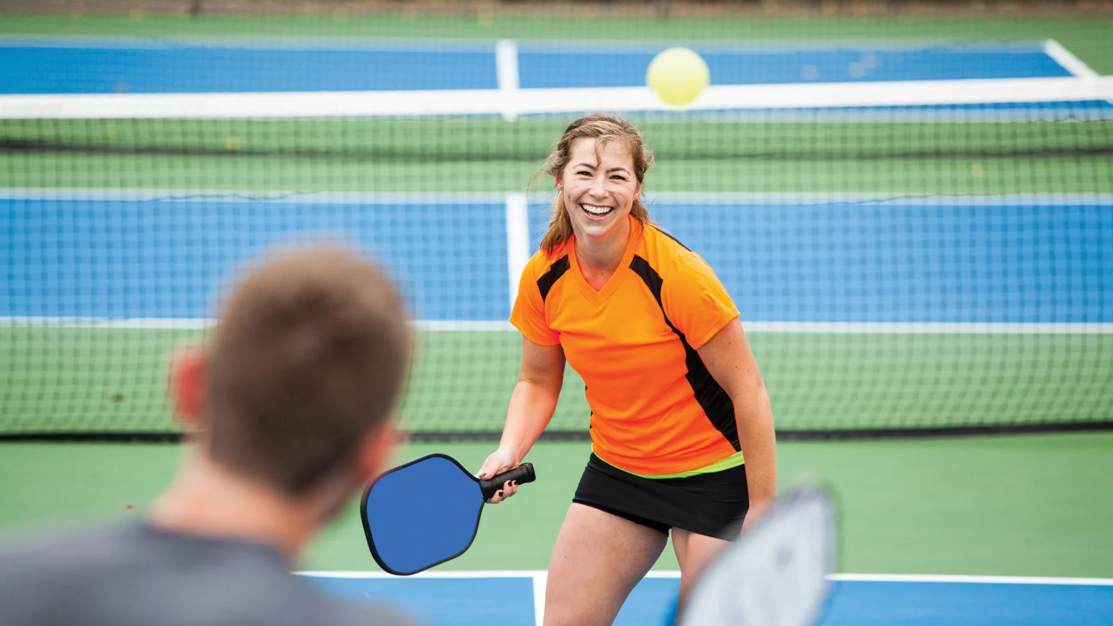 Girl on a pickleball court about to return the pickleball in a dink to her partner. She is wearing an orange and black athletic tee and tennis skirt and sneakers. New pet-friendly apartment homes for rent with private fenced backyards in Arizona, Texas, Florida, and North Carolina with pool, pickleball, dog park, gym, clubhouse and gated community. *No breed or weight restrictions. Yardly